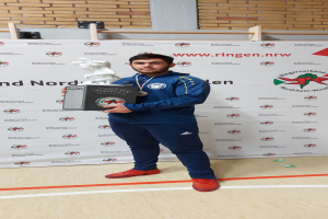 Ein Mann posiert stolz vor einer mit einem Banner geschmückten Wand, hält einen Pokal in der Hand und trägt eine Medaille um den Hals.