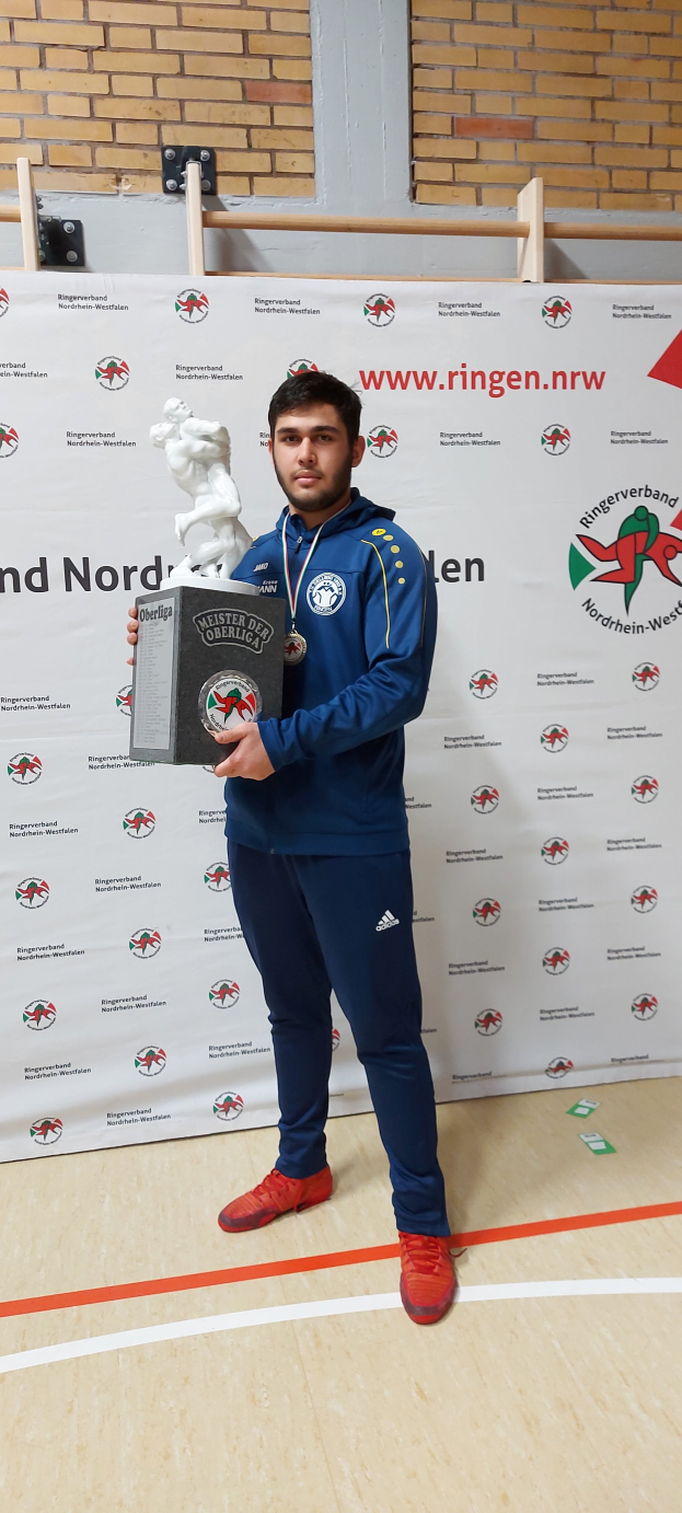 Ein Mann posiert stolz vor einer mit einem Banner geschmückten Wand, hält einen Pokal in der Hand und trägt eine Medaille um den Hals.