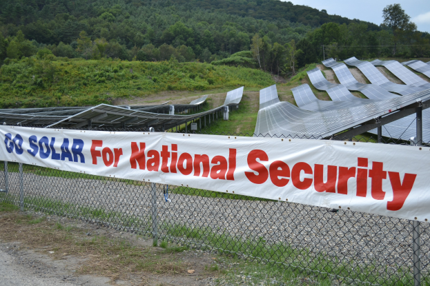 Ein Zaun mit einer 'Geht Solar für die nationale Sicherheit' -Aufschrift vor einem Solarpanel-Feld, Bäumen, Pflanzen und Gras unter einem klaren blauen Himmel.