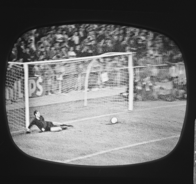 Ein Schwarz-Weiß-Foto eines Fußballspielers, der vor einem Tor auf dem Boden liegt, mit einem Ball in der Mitte und einem Netz im Hintergrund.