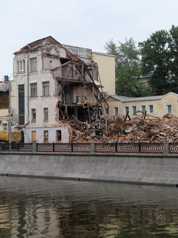 Teilweise abgerissenes Gebäude neben einem Fluss, mit einem Zaun und Bäumen im Hintergrund.