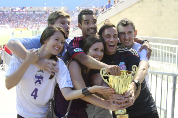 Sechs Menschen, zwei Frauen und vier Männer, die einen Pokal halten und lächeln, posieren für ein Foto vor einem Eisengeländer, mit einer Wand und einem Geländer oben rechts, Menschen und Lichter im Hintergrund, möglicherweise in einem Stadion.