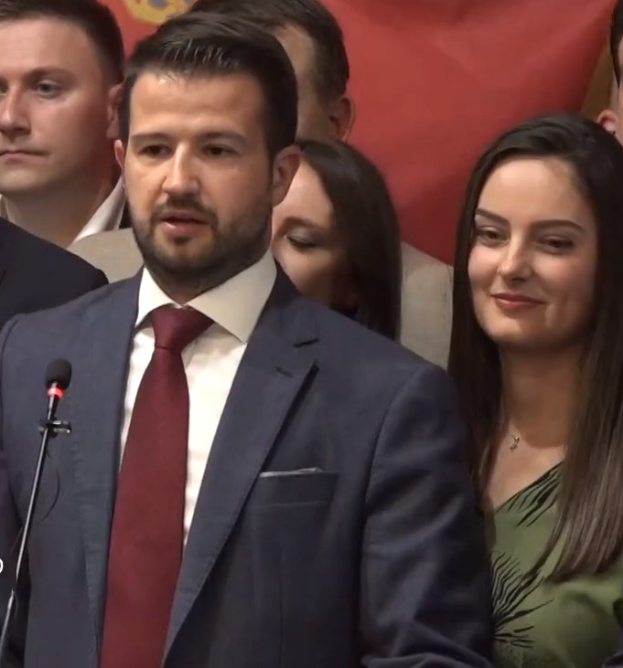 Ein Mann im Anzug spricht an einem Rednerpult mit einem Mikrofon, während er eine Menge anspricht und ein Banner im Hintergrund zu sehen ist.