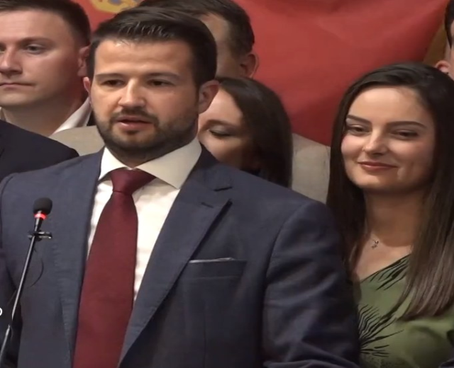 Ein Mann im Anzug spricht an einem Rednerpult mit einem Mikrofon, während er eine Menge anspricht und ein Banner im Hintergrund zu sehen ist.