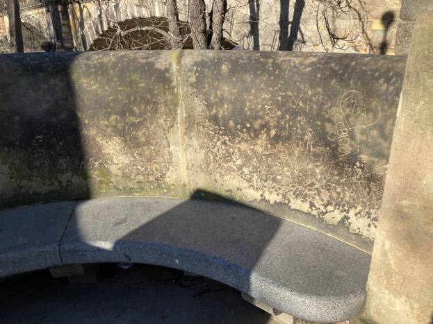 Eine alte Steinbank in einem Park, umgeben von einer Wand, Bäumen und einem Gebäude im Hintergrund.