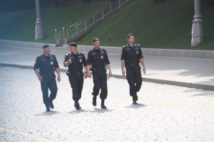Drei Polizisten in Uniform, die eine Straße entlanggehen, mit Pfosten, Geländern, Treppen und Gras im Hintergrund.