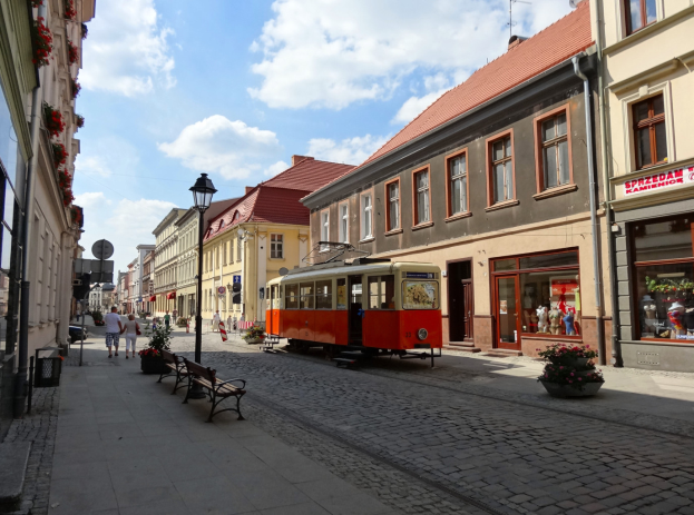 Eine rote und gelbe Straßenbahn fährt durch die Kopfsteinpflasterstraße der Altstadt von Tallinn, flankiert von Gebäuden mit Fenstern, Bänken und Laternen, mit Fußgängern auf den Gehwegen und einem bewölkten Himmel im Hintergrund.