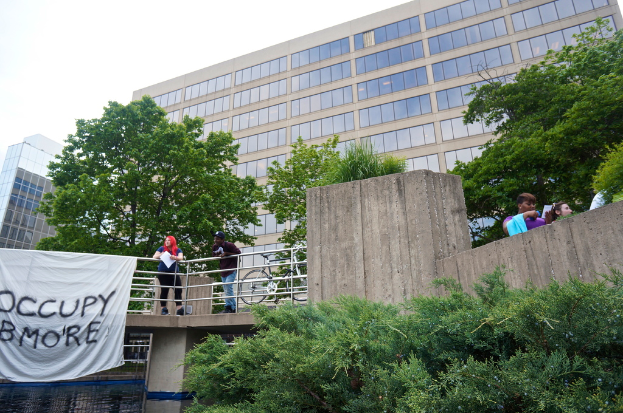 Menschen auf einer Brücke mit einem Banner und einem Fahrrad, unter der Wasser fließt, mit Bäumen, Gebäuden und Himmel im Hintergrund.
