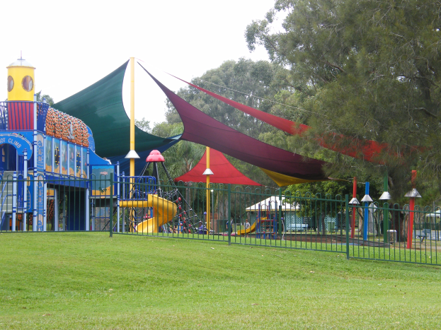 Ein Park mit einem Spielplatz, der von einem Metallzaun umgeben ist und mit einer Rutsche, Stangen und einem Zelt ausgestattet ist, auf dem Kinder auf saftig grünem Gras unter einem klaren blauen Himmel mit Bäumen im Hintergrund spielen.