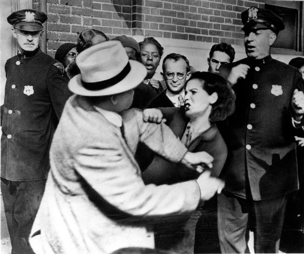 Zwei Personen kÃ¤mpfen in einem Schwarz-WeiÃŸ- Bild, mit mehreren anderen Personen im Hintergrund, einige tragen KÃ¤ppchen, und eine Wand dahinter.