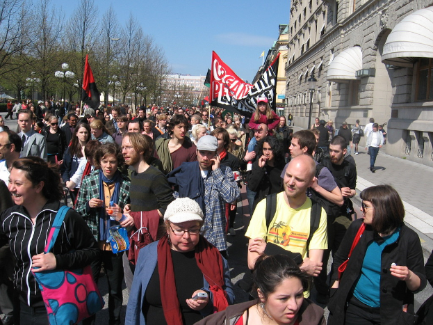 Personen im Vordergrund mit Gebäuden, Flaggen und einer Uhr an der Wand des zentralen Gebäudes im Hintergrund.