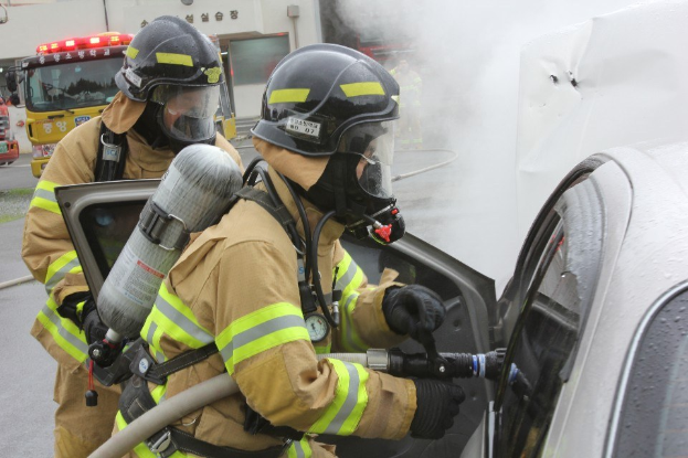 Feuerwehrleute in Schutzausrüstung löschen ein brennendes Auto mit einem Schlauch, während Rauch aus ihm aufsteigt, mit Fahrzeugen und einem Gebäude im Hintergrund.