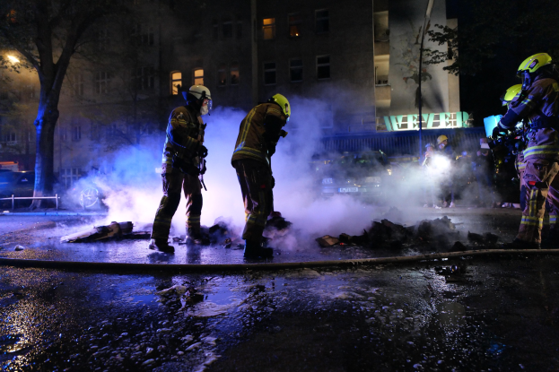 Feuerwehrleute in Helmen stehen nachts auf einer Straße mit Rauch aus einem Rohr, halten Gegenstände, während Gebäude, Bäume und Fahrzeuge im Hintergrund zu sehen sind, mit einer Tafel rechts.