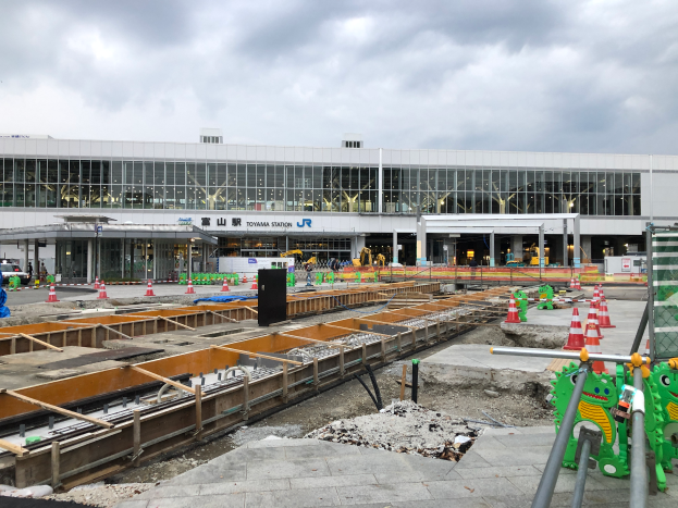 Baustelle am Toyama-Bahnhof mit einem großen Bauwerk im Hintergrund, umgeben von Fahrzeugen, Verkehrskegeln, Pfählen, Schildern und anderen Gegenständen, unter einem bewölkten Himmel, mit einem Wasserzeichen unten.