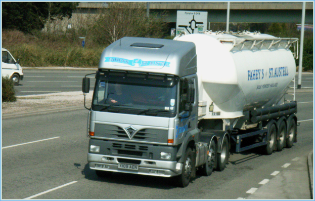 Ein Lastwagen mit Fahrer Ã¼berquert eine StraÃe, mit einem Van auf der Gegenfahrbahn, BÃ¤umen und einer BrÃ¼cke im Hintergrund.