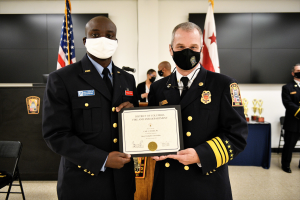 Zwei uniformierte Männer mit Masken halten eine Urkunde neben einem Rednerpult mit Mikrofon, einem Stuhl links daneben, Flaggen im Hintergrund und Trophäen auf einem Tisch davor.