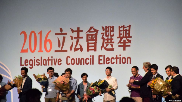 Eine Gruppe von Menschen auf einer Bühne mit Blumensträußen in den Händen, mit einem Publikum darunter und einem Bildschirm mit Text und Logo für die Wahl des Legislativrats 2016 im Hintergrund.