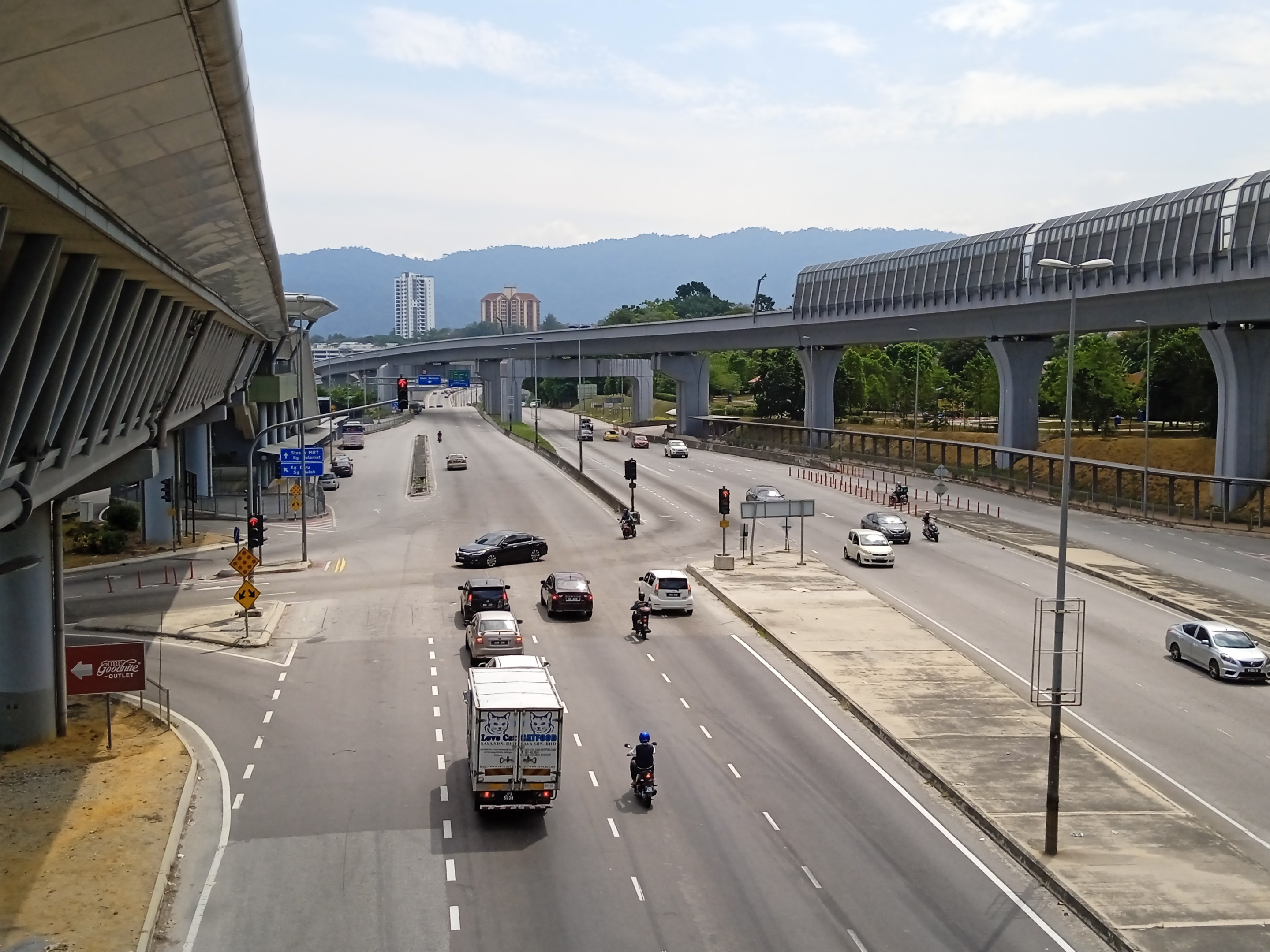Eine vielbefahrene Autobahn mit mehreren Fahrspuren neben einer Brücke, mit Straßenlaternen, Verkehrsampeln, Verkehrsschildern, Bäumen, Gebäuden, Hügeln und einem bewölkten Himmel.