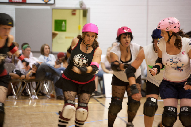 Frauen in Helmen und Knieschonern spielen einen Sport, mit einer Gruppe von Menschen im Hintergrund, einer Wand, einer Tür und Brettern oben und einem Boden unten.