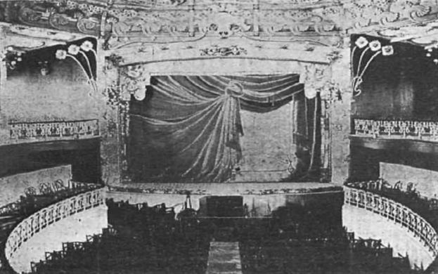 Schwarzes und weißes Foto des Auditoriums des Theatre Royal in London, das eine Bühne mit Vorhang, Geländer, verzierte Wände und sitzende Zuschauer zeigt, mit Text unten.