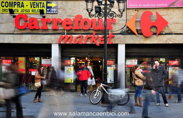 Ein belebter Einkaufsmarkt mit Menschen vor einem Einkaufszentrum, einem schwarzen Pfahl mit Lichtern, einem Fahrrad daneben und einem Richtungsschild an der Wand.