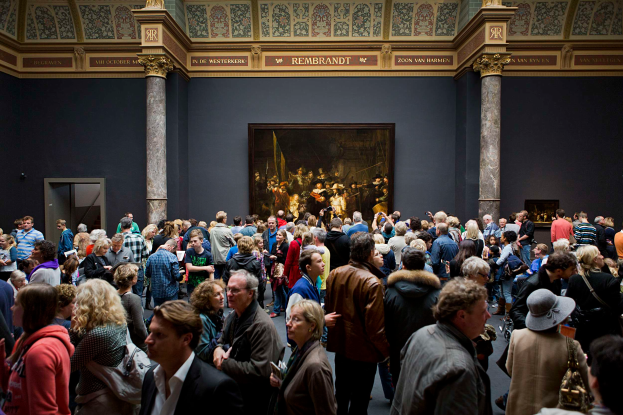 Eine große Gruppe von Menschen in einem Raum mit einem Foto auf der Wand, Säulen, einer Tür und einer Decke mit Lampen, einige halten Taschen, im Museum of Fine Arts in Amsterdam.