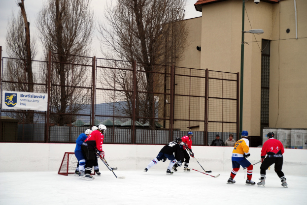 Personen spielen Eishockey auf einer Eisfläche mit Gebäuden, Bäumen, einer Straßenlaterne, einem Namensschild und Zäunen im Hintergrund bei klarem Himmel.