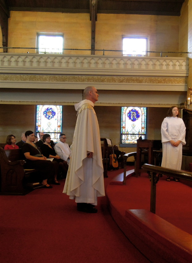 Ein roter Teppich führt zu einer Szene, die wie eine Kirche aussieht, mit einem Mann und einer Frau in weißen Kleidern, die in der Nähe eines hölzernen Geländers und einer Kerze stehen, Menschen auf Bänken sitzen und Musikinstrumente im Hintergrund.