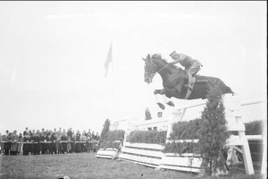 Schwarz-weiß-Foto eines Pferdes und Reiters, die über ein Hindernis springen, bei den Royal Ascot Horse Trials 1953, mit Zuschauern, einer Fahne und Grün im Hintergrund.