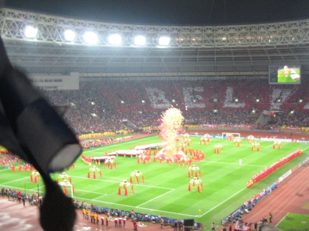 Ein Stadion mit Flutlicht, Eisengittern, Zuschauern, stehenden Personen, Werbetafeln, Anzeigetafeln, ├ťberwachungskamera und verschiedenen Fahnen und Deko.