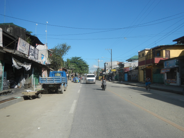 Eine städtische Straße mit fahrenden Fahrzeugen, ein Radfahrer auf der rechten Seite, Gebäude auf beiden Seiten, Bäume im Hintergrund und ein klarer blauer Himmel.
