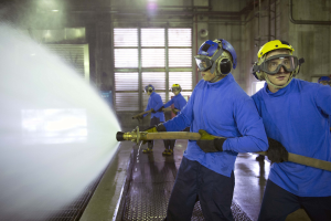 Gruppe von Männern in blauen Hemden und gelben Helmen bei der Arbeit an Maschinen, einer davon sprüht Wasser auf den Boden in einer Fabrikumgebung.