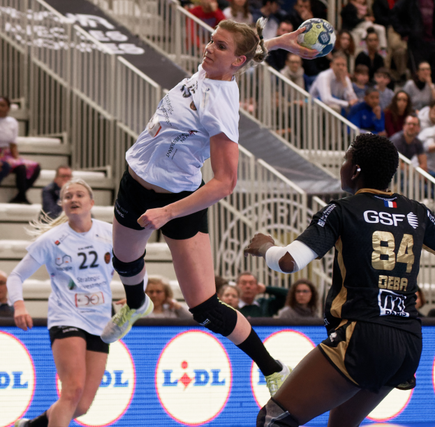 Eine Gruppe von Frauen, die auf einem Handballfeld spielen, mit einem Ball in der Luft, Zuschauern auf Treppen und Geländern im Hintergrund und einem Schild mit der Aufschrift "Handball-Weltmeisterschaft 2019 - Tag 2".