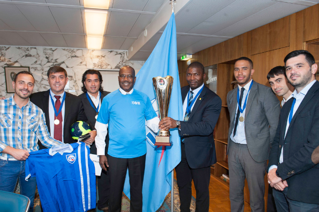 Eine Gruppe von Menschen, die zusammen posen, wobei zwei in der Mitte einen Pokal halten, einer links ein T-Shirt und ein anderer links einen Ball hält, und eine Flagge im Hintergrund.