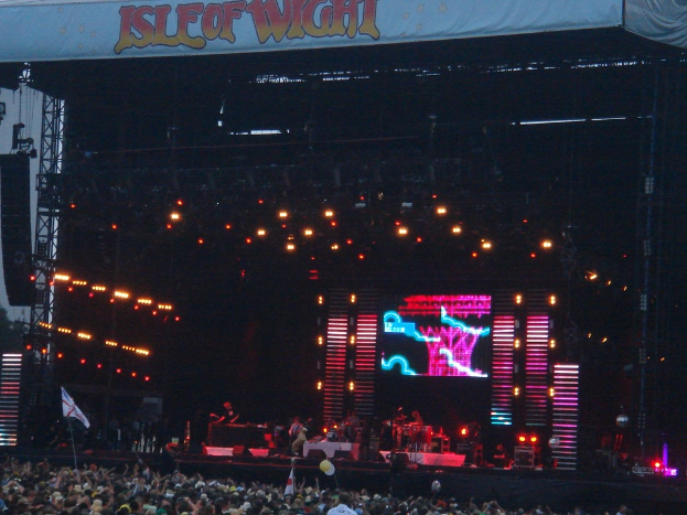 Eine Bühne mit Lichtern, einem Bildschirm und Musikern, die Instrumente spielen, mit einem Banner oben, einer Menge unten und Fahnen mit Stangen und Ballons drum herum.