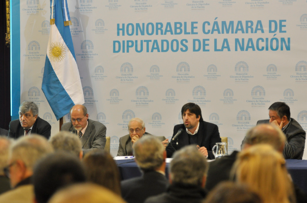 Eine Gruppe von Menschen sitzt in Reihen von Stühlen, vor der ersten Reihe steht ein Tisch mit einem Krug, Papieren und einem Stift. Eine Person in der Mitte spricht in ein Mikrofon, hinter ihnen eine weiße und blaue Flagge und ein weißer und blauer Hintergrund mit der Aufschrift "ehrenwerte kamara de".