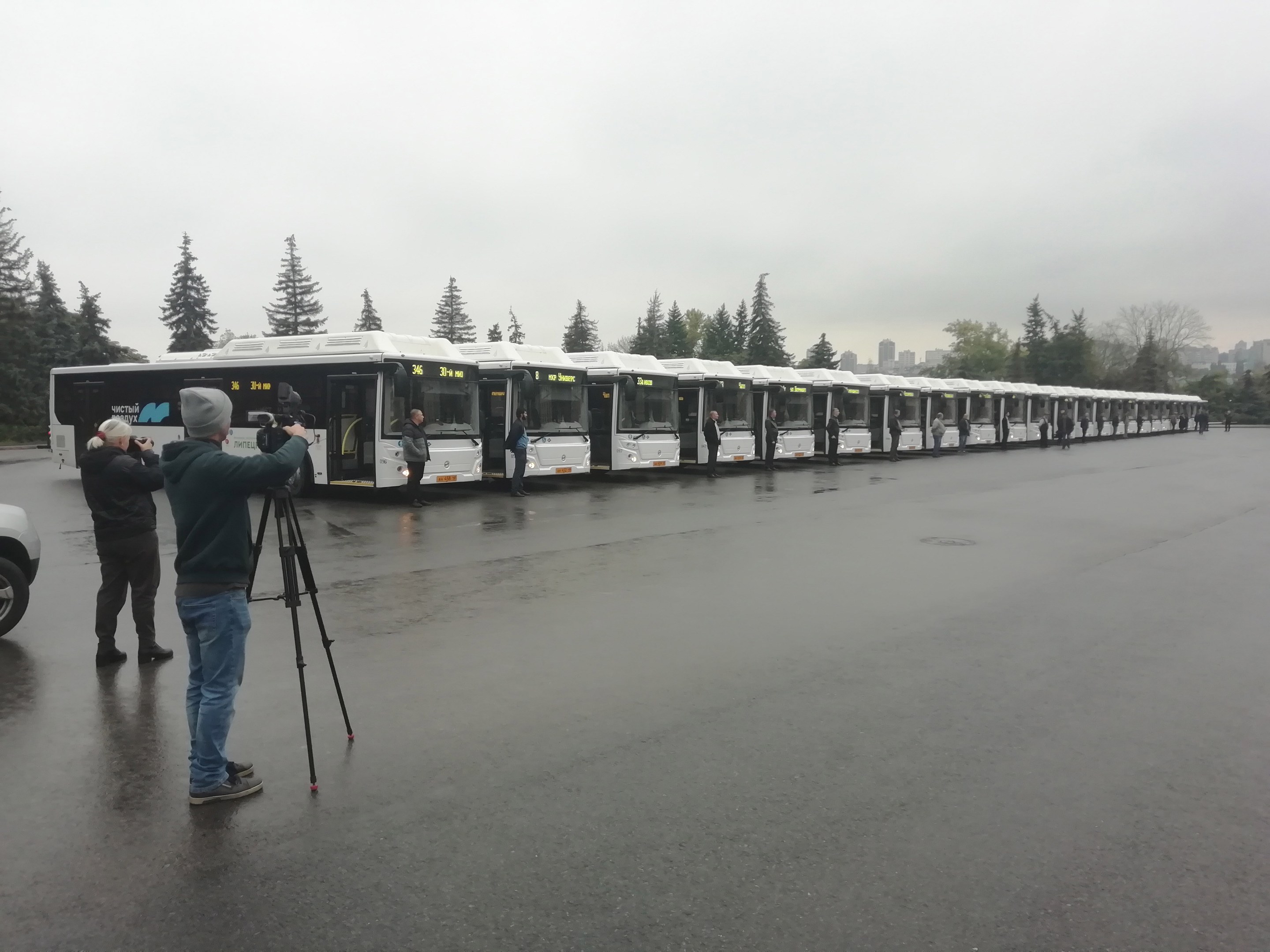 Eine Gruppe von Menschen, die eine Reihe von Bussen auf einer Straße mit Bäumen, Gebäuden und einem klaren blauen Himmel fotografieren.
