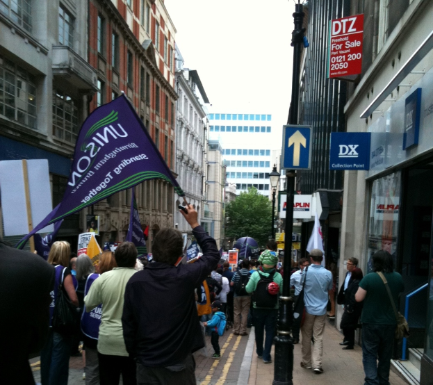 Eine große Gruppe von Menschen auf einer Straße, einige halten Fahnen und Plakate, mit einem Schild an einem Pfahl, Gebäuden mit Fenstern an den Seiten und einem Baum, Laternenmast und Gebäude im Hintergrund unter einem Himmel.