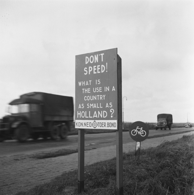 Schwarz-weißes Foto eines Lkws auf einer Straße mit einem "Fahren Sie nicht zu schnell"-Schild im Vordergrund und anderen Fahrzeugen, Gras, Masten und einem klaren Himmel im Hintergrund.