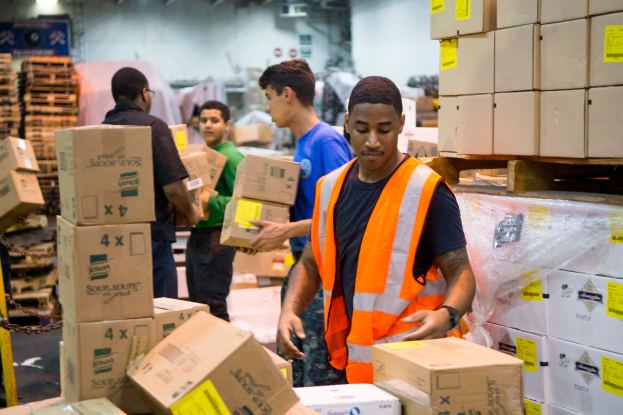 Eine Gruppe von Männern in orangefarbenen Westen und Sicherheitswesten lädt Kisten in ein Lagerhaus, mit mehreren Kartons auf der rechten Seite und einer Kette auf der linken Seite, und einer Wand mit Lampen im Hintergrund.