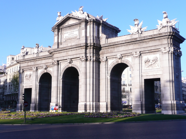 Eine Szene mit Gebäuden und Skulpturen in der Mitte, einer Straße, Gras, Blumen, einem Pfahl und einer Tafel unten und einem Himmel oben.