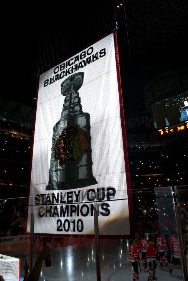 Eine nächtliche Stadion-Szene mit einem großen Banner in der Mitte, Zuschauern im Hintergrund, einem Glas auf dem Boden, einigen Spielern auf dem Feld und beleuchteten Tribünen.