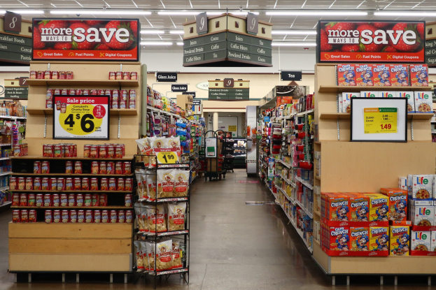 Ein Kiosk-Innenraum Mit Verschiedenen Lebensmitteln, Die Auf Regalen, Regalen Und Dem Boden Angezeigt Werden, Einschl. Schilder Mit Text Und Zahlen, Deckenbeleuchtung Und Einem Menschen Im Hintergrund.