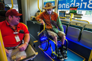 Zwei Männer in Rollstühlen in einem Bus, einer trägt ein rotes T-Shirt und eine rote Mütze, der andere ein orangefarbenes Hemd und einen braunen Hut, beide tragen Masken und Ausweise, mit gelben Stangen und Glasfenstern, die Bäume, Gebäude und den Himmel zeigen.