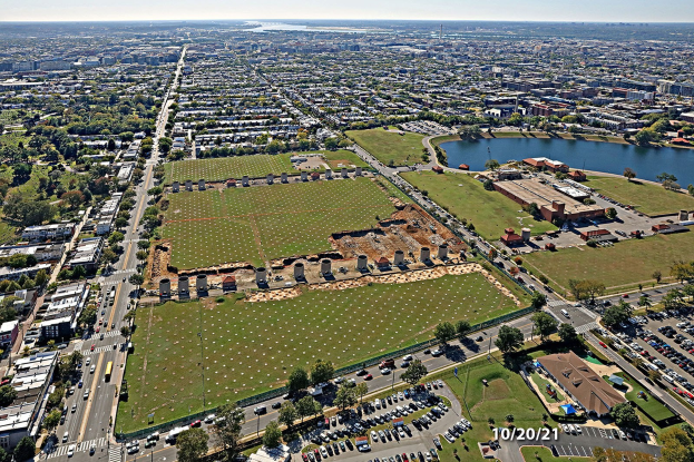 Luftaufnahme einer Stadt mit Gebäuden, Bäumen, Gras, Straßen mit Fahrzeugen, Wasser und Wolken, die einen großen zentralen Platz als Park zeigen.
