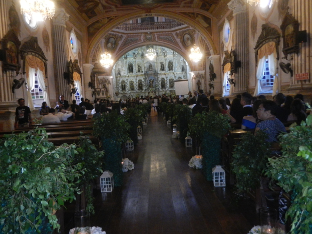 Eine Hochzeitszeremonie in einer Kirche mit Gästen auf Bänken sitzend und auf dem Boden stehend, Topfpflanzen schmücken den Gang, Skulpturen, Vorhänge an den Fenstern und Kronleuchter an den Wänden, sowie ein Bildschirm im Hintergrund.