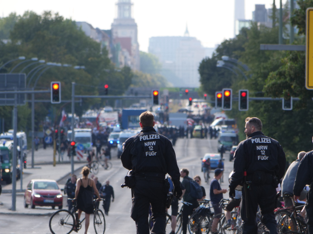 Polizeibeamte fahren mit Fahrrädern eine von Bäumen gesäumte Straße entlang mit Gebäuden und einem klaren Himmel im Hintergrund.