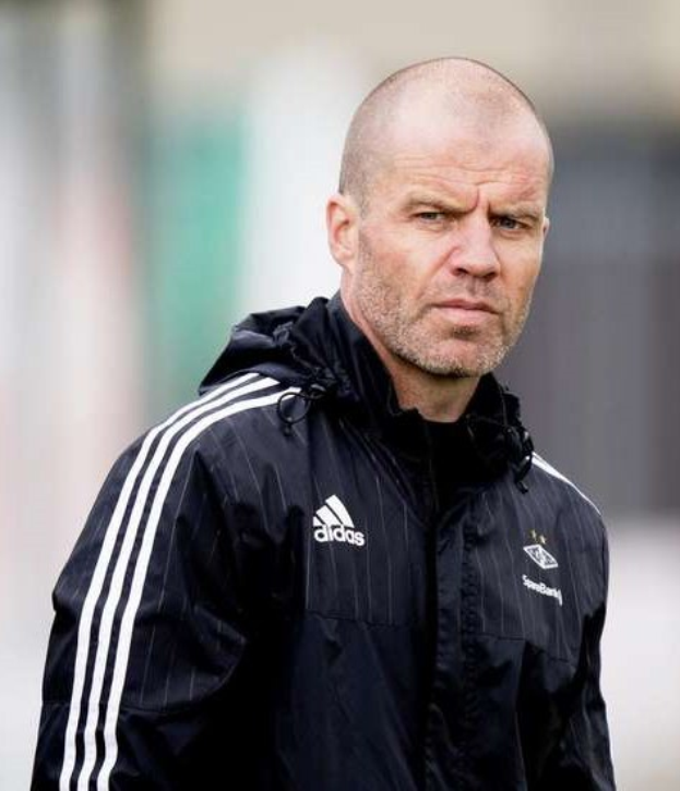 Baldiger Trainer in einer marineblauen Jacke mit weißen Streifen und weißem Kragen, der auf einem Fußballfeld steht und die Hände verschränkt hat.