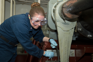 Eine Frau in blauer Uniform und Schutzbrille arbeitet an einer Maschine, umgeben von Rohren und einer Wand im Hintergrund.