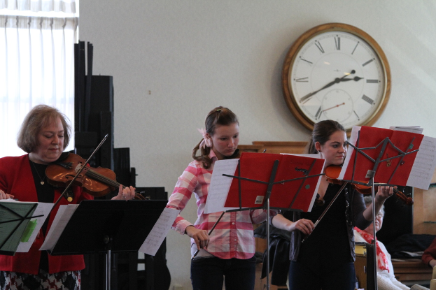 Drei Frauen auf der Bühne mit Geigen stehend und Notenständer vor sich, während eine Gruppe von sitzenden Frauen im Vordergrund zuhört; eine Uhr hängt an der weißen Wand im Hintergrund, mit einem weißen Vorhang in der linken Ecke.